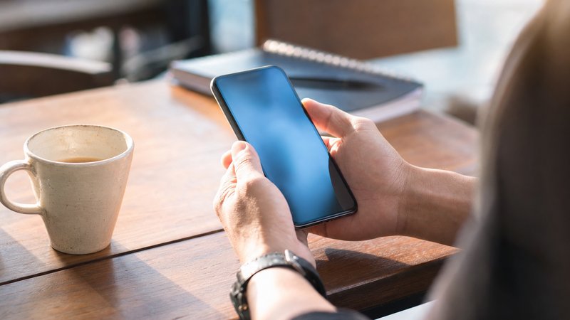 A person checking a phone at a sunlit cafe table with a coffee cup and notebook beside them