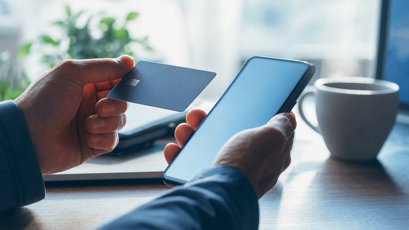 A person holding a plain bank card over a smartphone at a sunlit desk