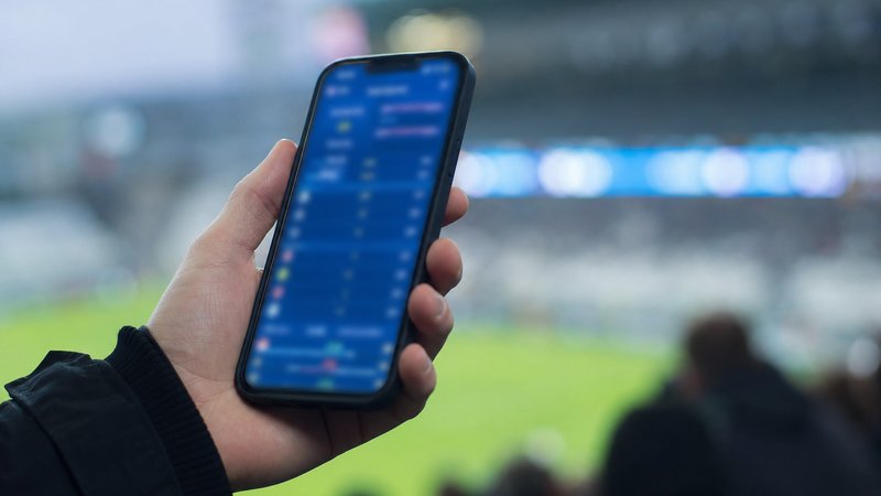 A hand holding a phone showing a betting app while watching a match at a stadium