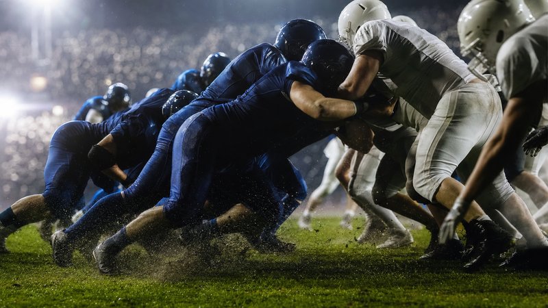 An American football on the yard line of a floodlit stadium under evening lights